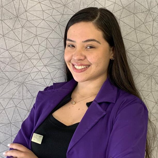 Secretária Larissa Amorim vestida com terninho roxo pousando de braços cruzados e sorrindo para a foto