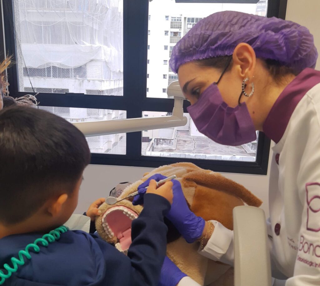 Odontopediatra de touca e mascara lilás mostrando um modelo de boca para uma criança de roupa azul durante uma consulta