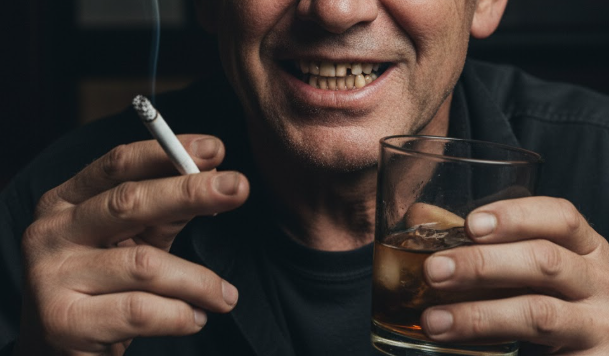 Homem com cigarro na mao direita, bebida na mao esquerda e dentes amarelados